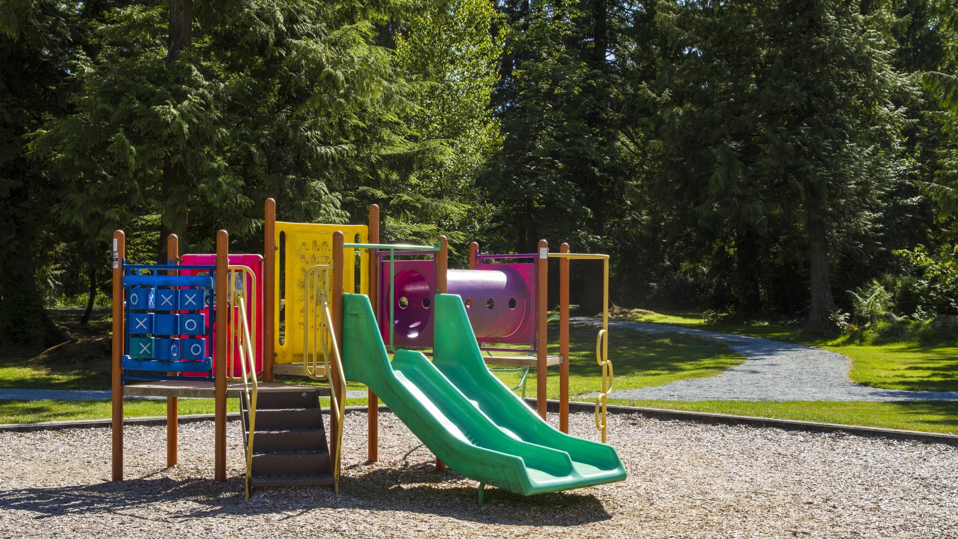 A small, colourful playground. Beyond it, a wide walkway leads into a forested area.